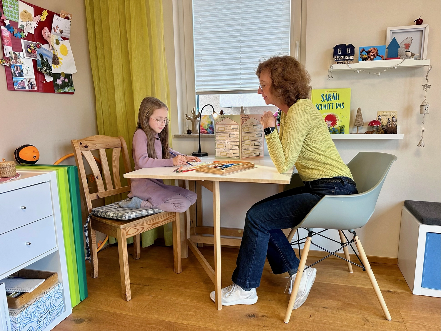 Zeit & Raum für Sie und Ihr Kind – Erzieherin und Heilpädagogin – Dieses Bild zeigt eine Frau und ein Mädchen, die an einem kleinen Tisch in einem Raum sitzen, möglicherweise für eine Lernsitzung oder Aktivität. Der Raum ist mit verschiedenen Gegenständen dekoriert, darunter einem Buch mit dem Titel „Sarah schafft das“.